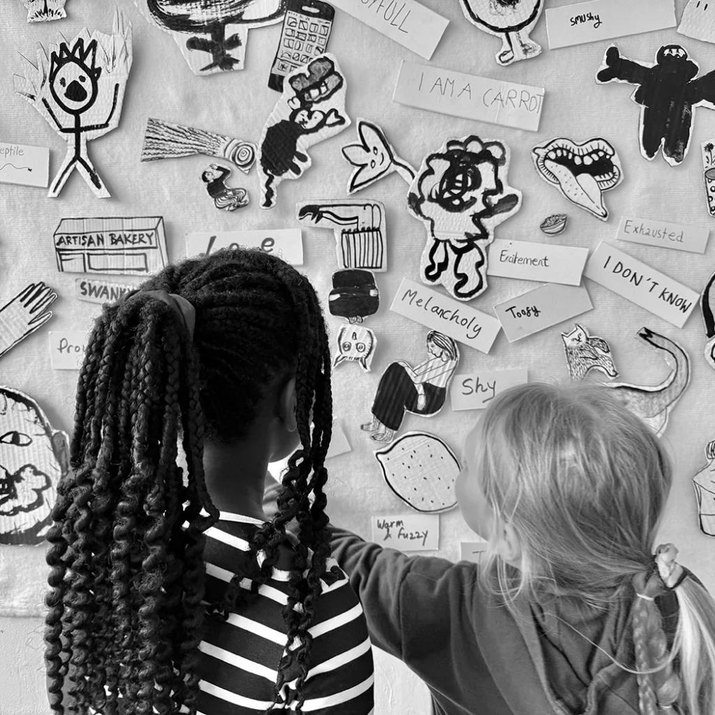 Two children with different hairstyles examine a wall filled with hand-drawn doodles and emotion labels in black and white.