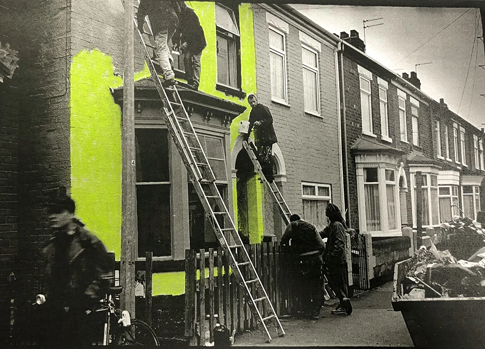 Black and white photo of workers on ladders painting bright neon yellow sections on residential brick houses