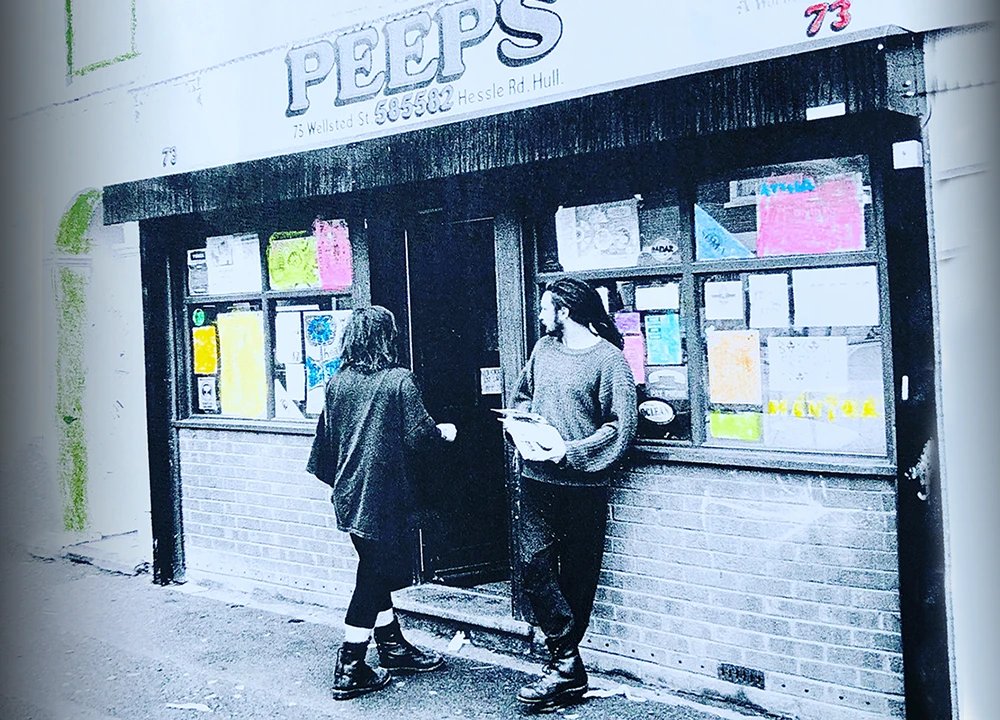 Two people standing outside Peeps shop with colorful posters, black and white photo with color accents