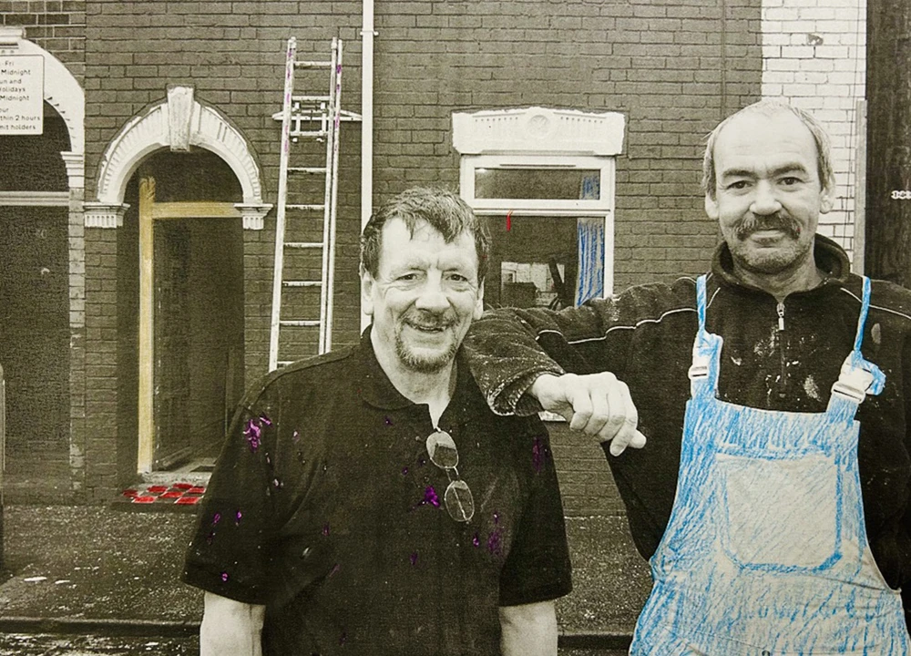 Two painters in work attire smiling outside brick building with ladder, wearing splattered black and blue overalls